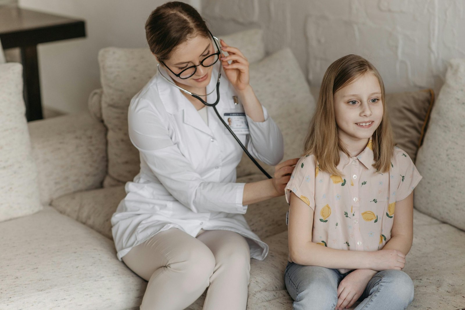 Doctor using a stethoscope for a child's check-up on a sofa during a home visit.